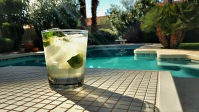 Picture of a cocktail with clear liquor, a lime and muddled mint with ice in front of a pool beautiful pool.  Looks like a summertime delight.  