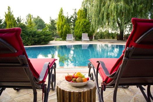 Picture of two red pool chair facing a pool with two more chairs facing from other side.  Pool is surrounded by beautiful foliage and luscious greens.