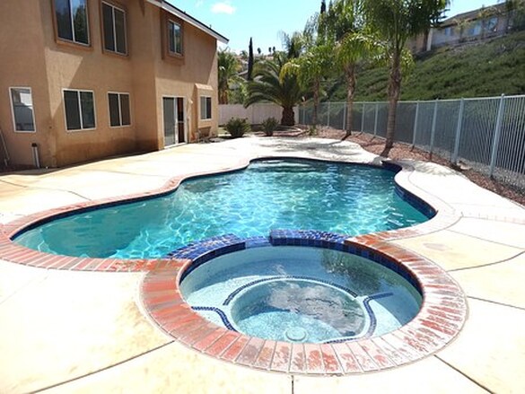 Picture of a crystal clear spa in front of a pool with brick lining and tile floors.  Behind are palm trees, fences, and residential houses.